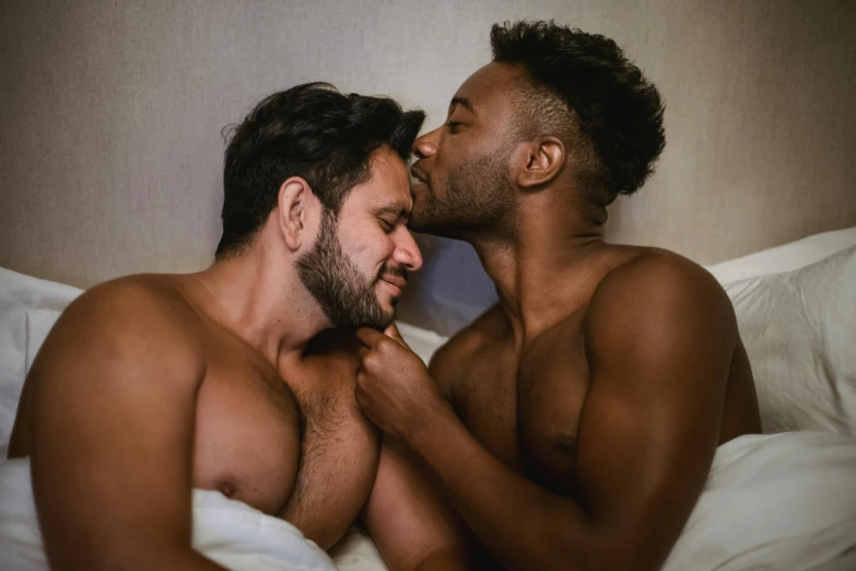 two men sitting in bed one holding the other's face and kissing