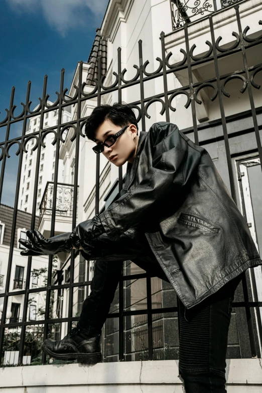 a man wearing sunglasses, black jacket, and black boots leaning on a black fence