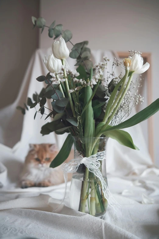 white flowers and greenery in a vase with a cat lying beside