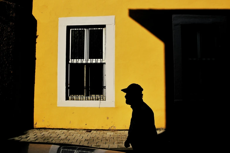 a man in the shadows walking past a yellow building