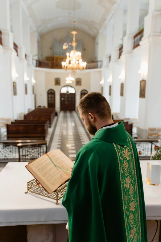 a young man in a green priest robe reading an open book in an empty building