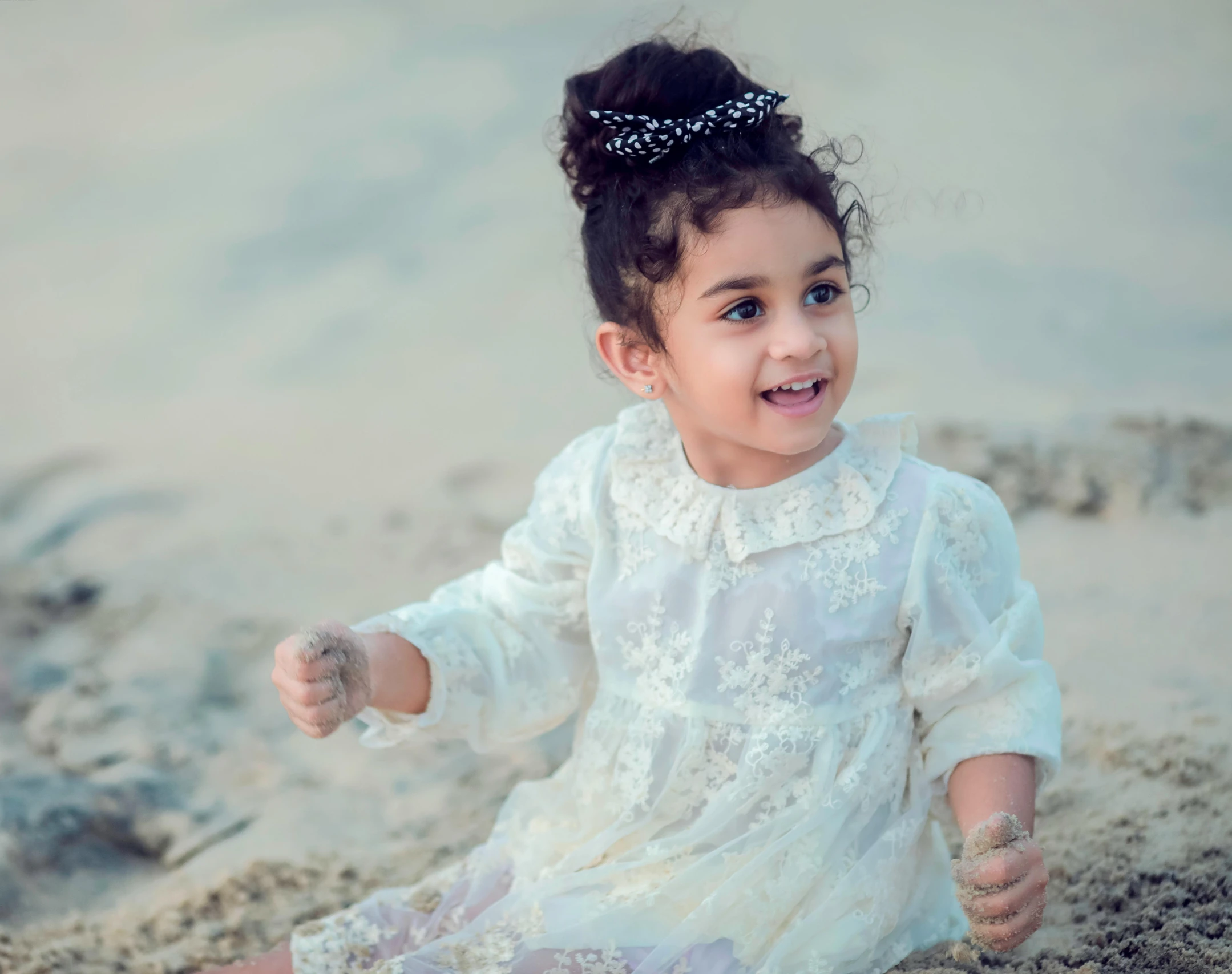 little girl with a tiara and pearls on her head, sitting on sand and wearing a white dress