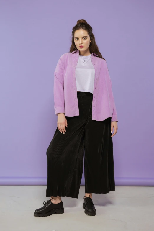 a  in a pink cardigan and black wide legged pants standing against a purple wall