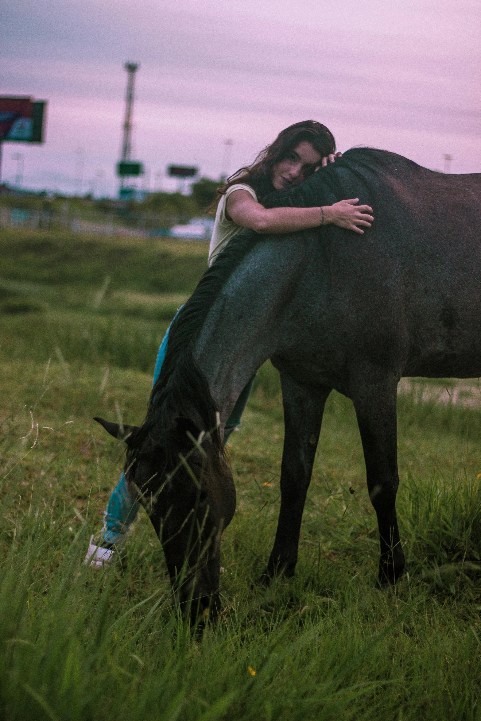 a woman hugs her horse while grazing in the green grass