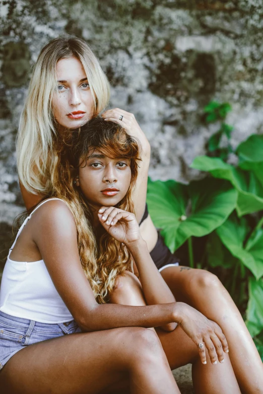 two women sit close to each other by some bushes