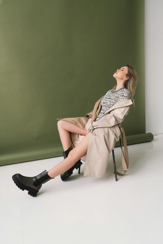 a woman in short shorts, long coat, and black boots sits against a green wall