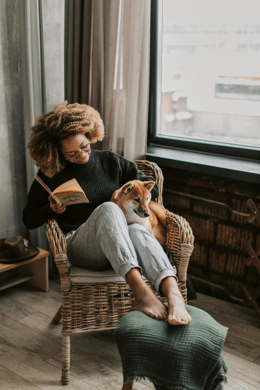 a woman sitting in a wicker chair reading with a dog sitting next to her
