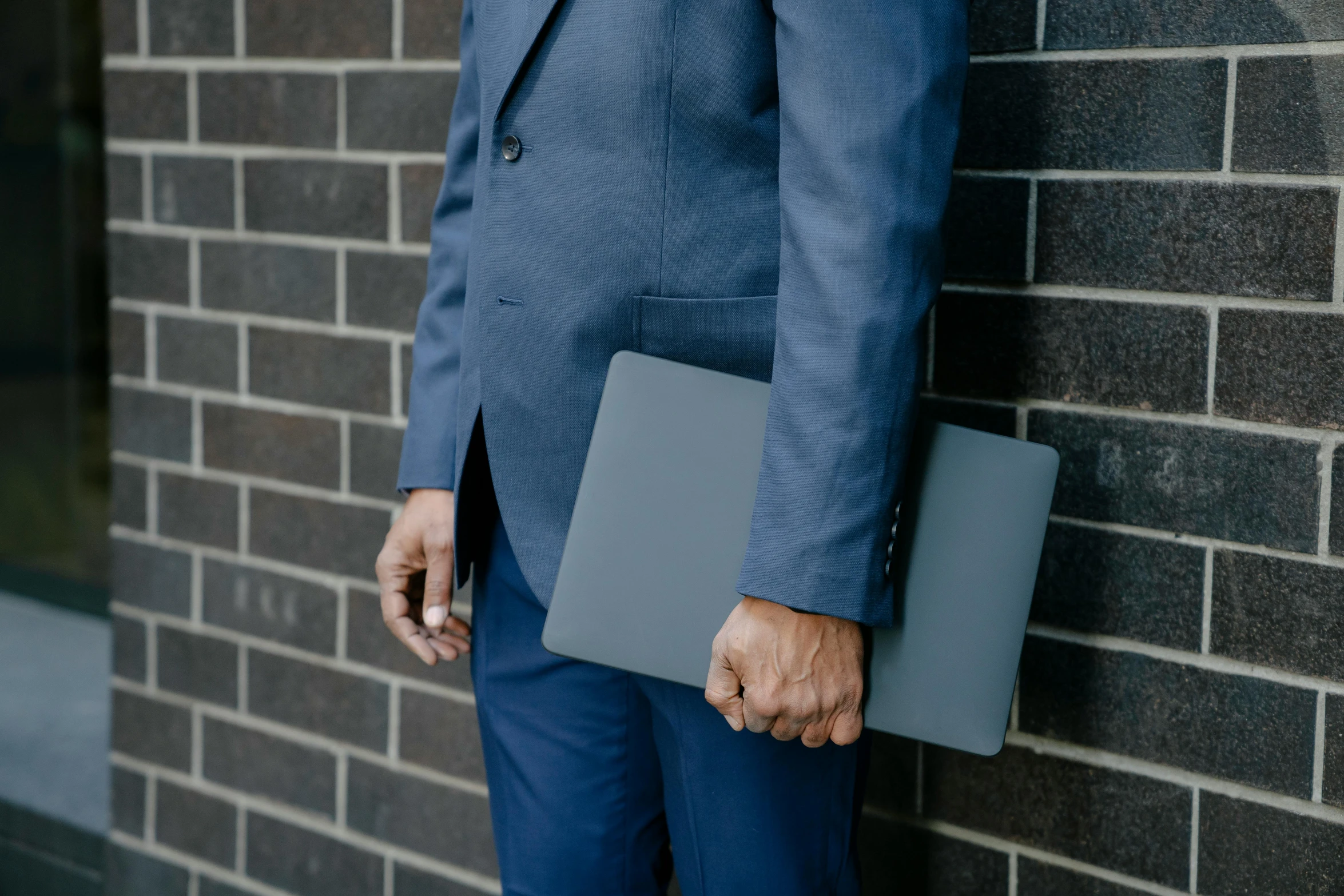 the suit man stands against a brick wall holding his laptop