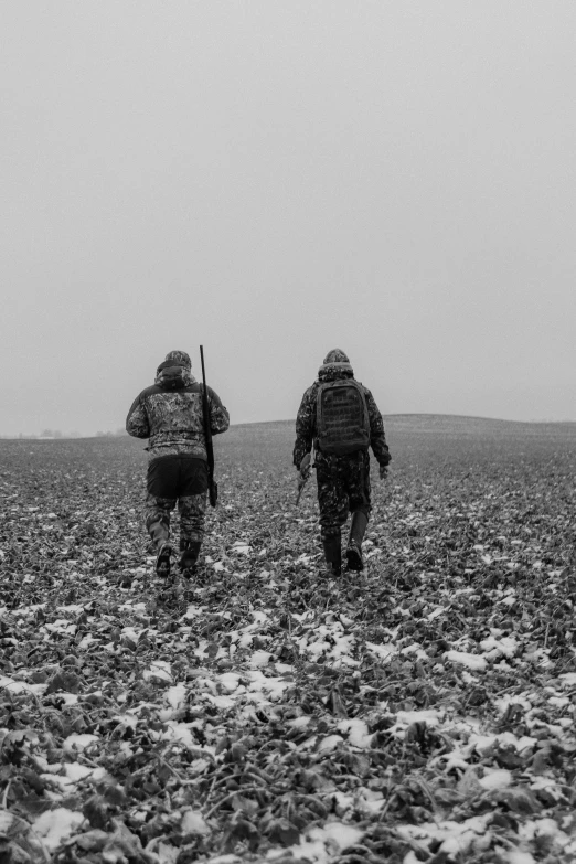 two men with guns walking away from the camera in a field