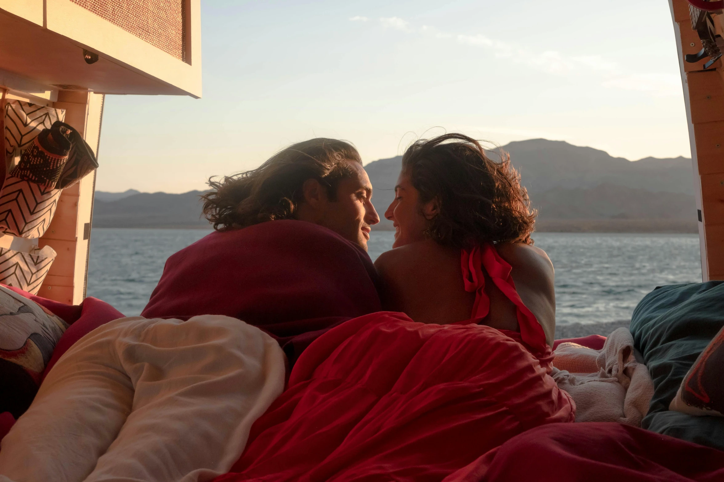 two people in bed looking at each other and sitting beside a lake