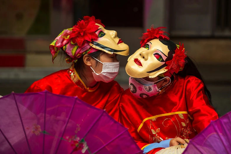 two people wearing red and yellow masks and holding pink umbrellas