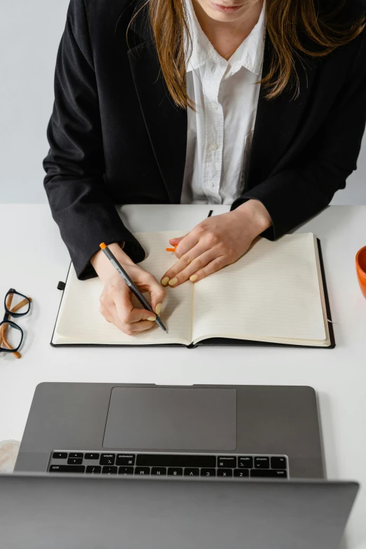 a woman in black jacket writing on book next to laptop