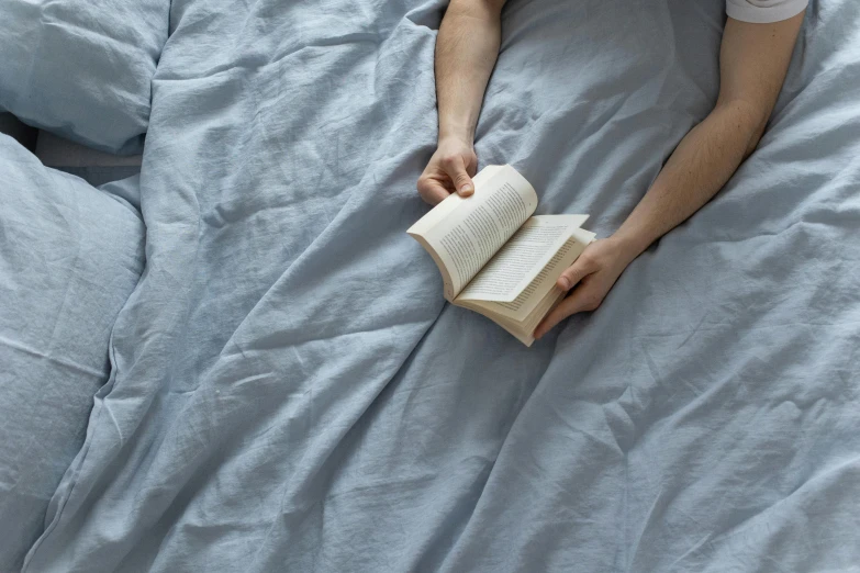 a person lays in bed with blue sheets and reads a book