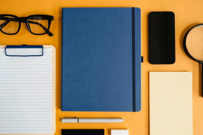 various items laid out on a surface to include a notebook, pen, glasses, pencil and eyeglasses