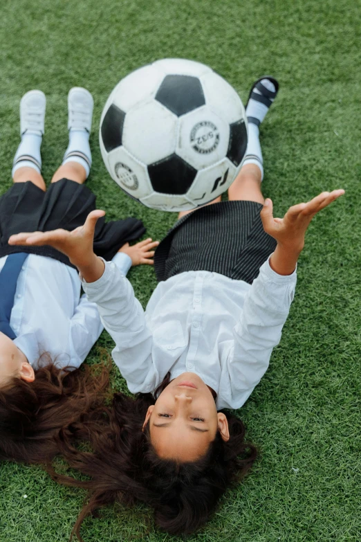 two children are on grass with their arms out as they lay next to a soccer ball