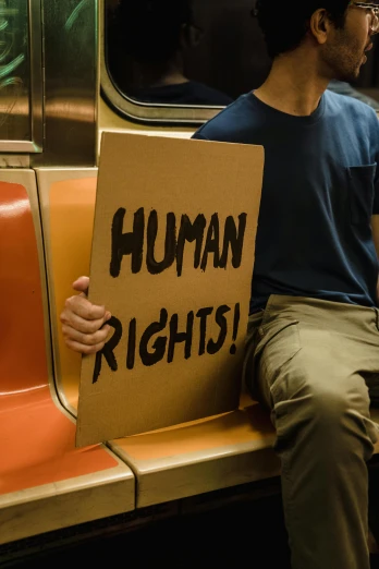 a man in glasses sits on a train while holding a sign that says human rights