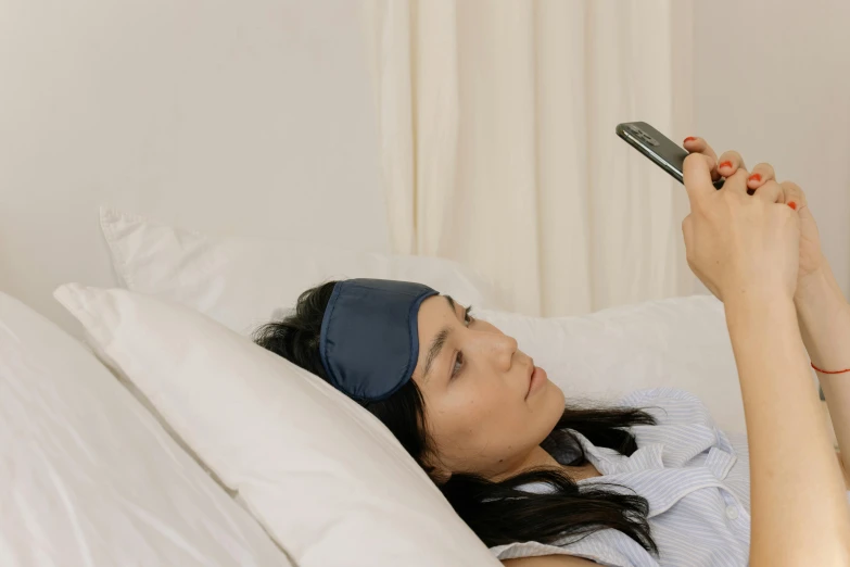 a young woman laying in bed with a sleeping headband on, holding up her cell phone