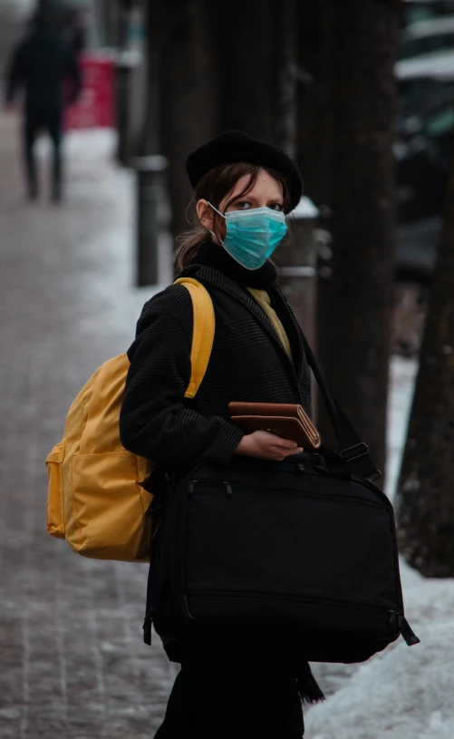 woman in a black jacket wearing a blue face mask carrying a suitcase