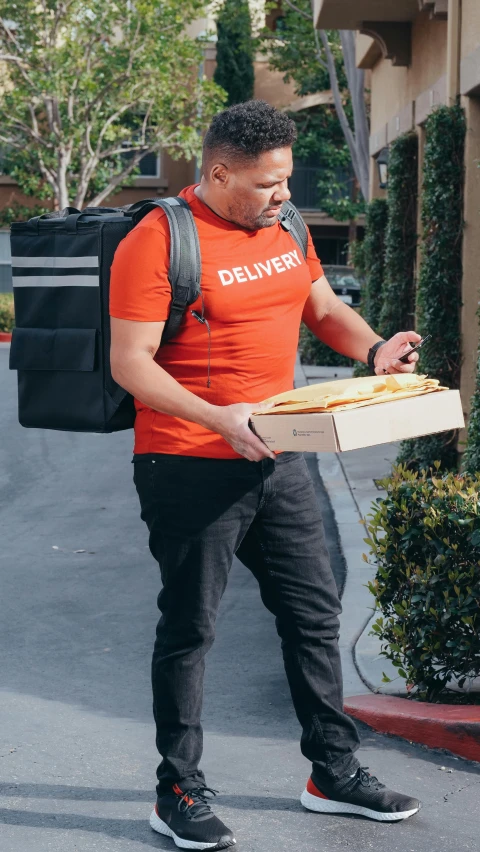 a man with an orange shirt carries a pizza in a black bag