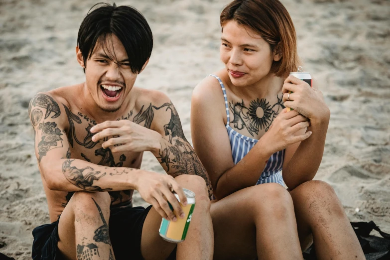 two people sit on the beach, one smiling and laughing