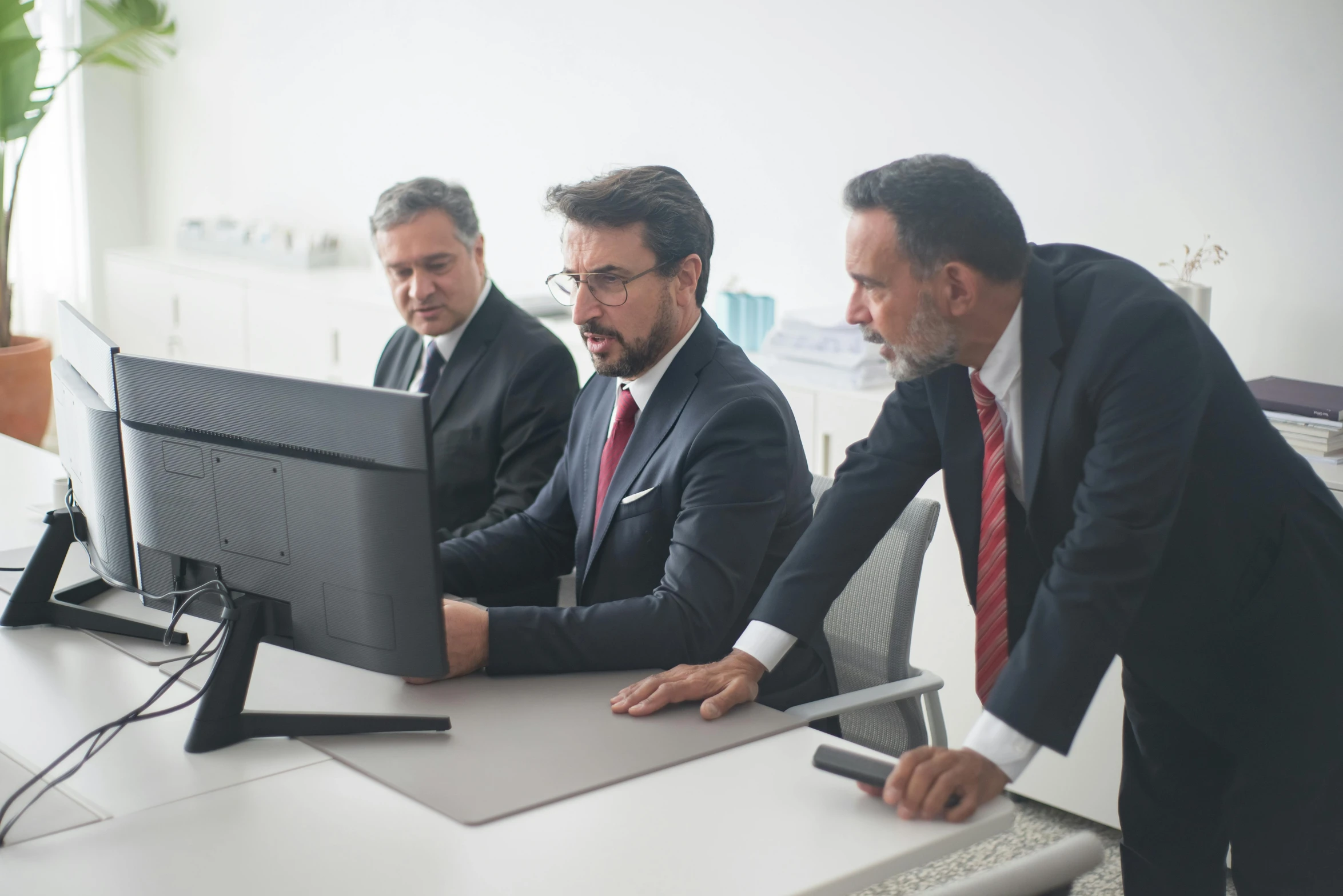 three men look at a computer monitor and laptop