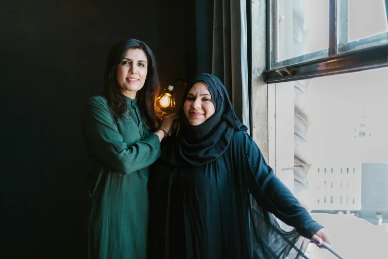 two woman in hijab standing next to each other in front of a window