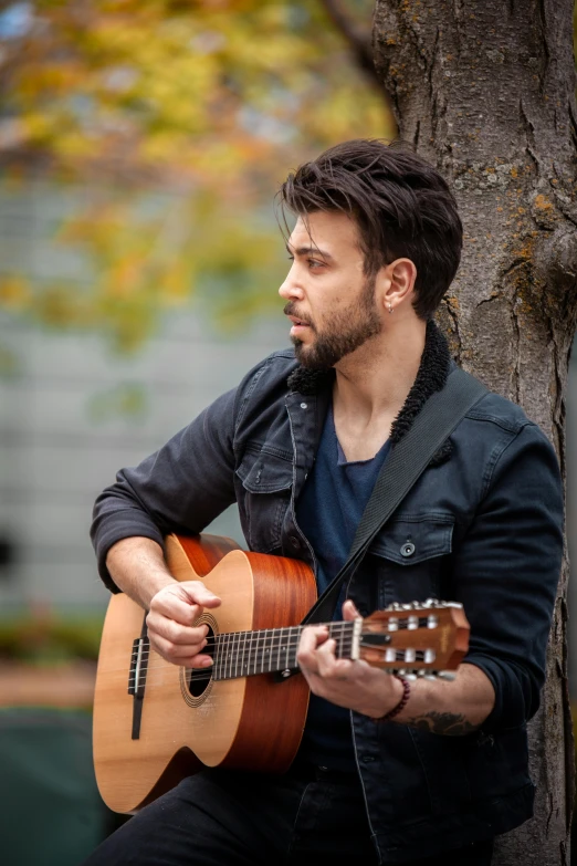 a man sitting next to a tree playing an acoustic guitar