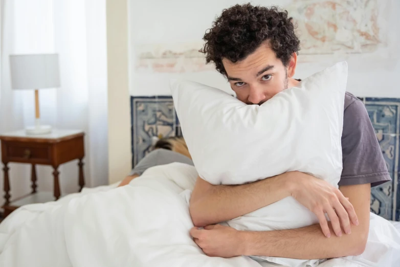 a man with curly hair laying in bed with a pillow over his head