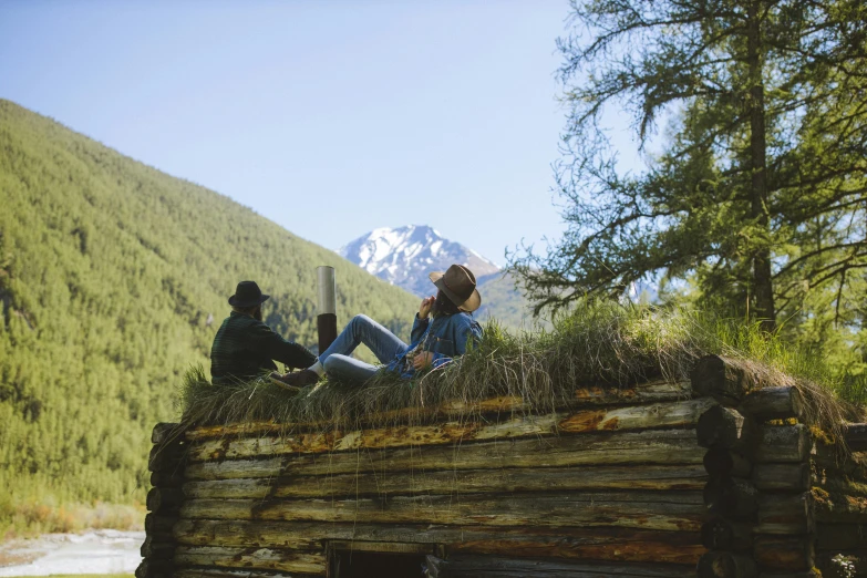 three people sitting on top of a log cabin, in front of a mountain