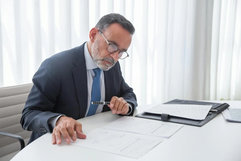 a man wearing a suit and tie with eye glasses, holding a pen, looking at some paperwork