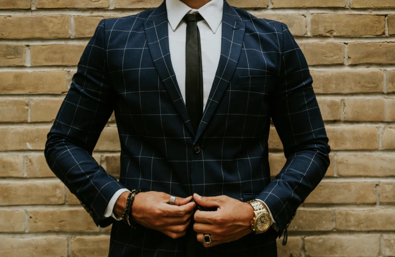 a man in a plaid suit and gold watch stands against a brick wall