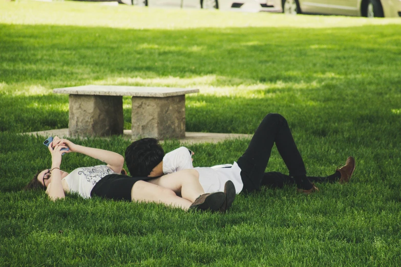 two people lie on the grass under a stone bench