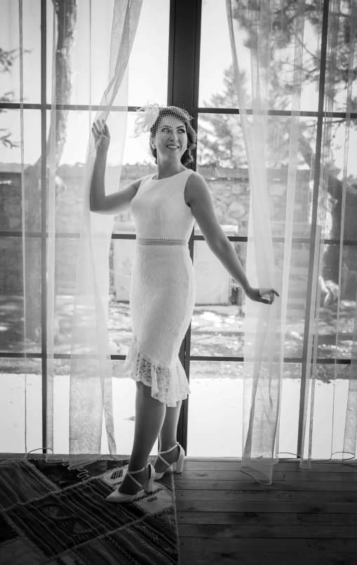 woman in white dress looking out window with sheer curtain