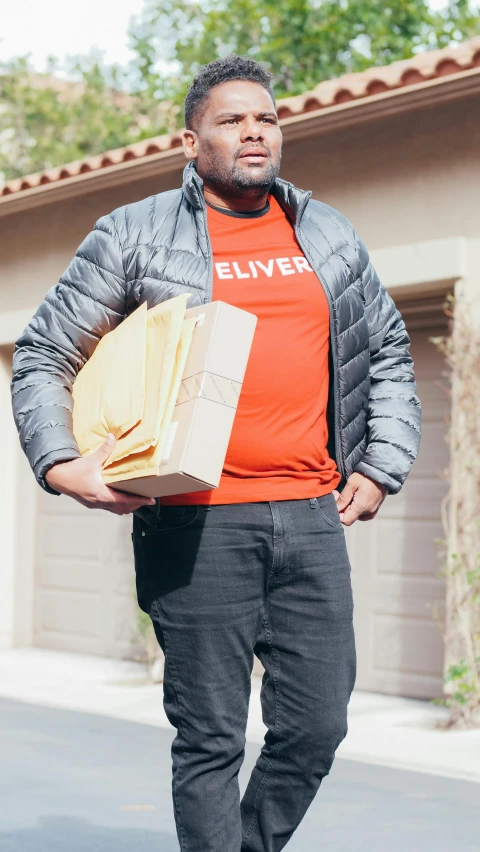 the man in orange shirt has his hands on his hips and is carrying two cardboard boxes in one hand