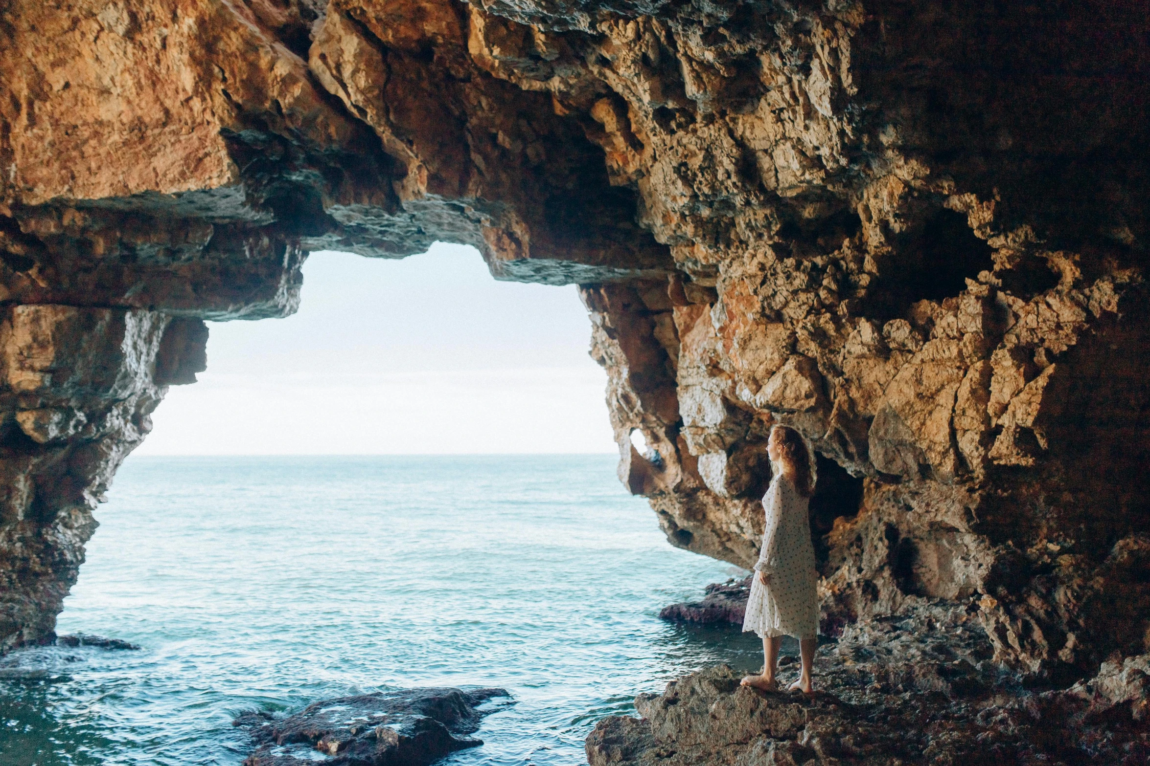 a woman in white clothing standing by large cave on water