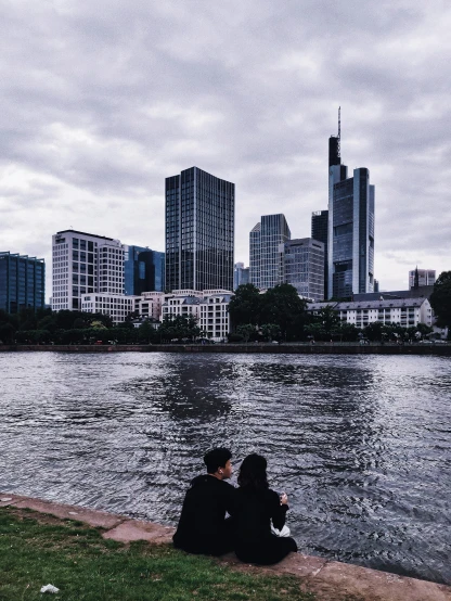 two people sitting on the bank of a river looking at the city in front of them