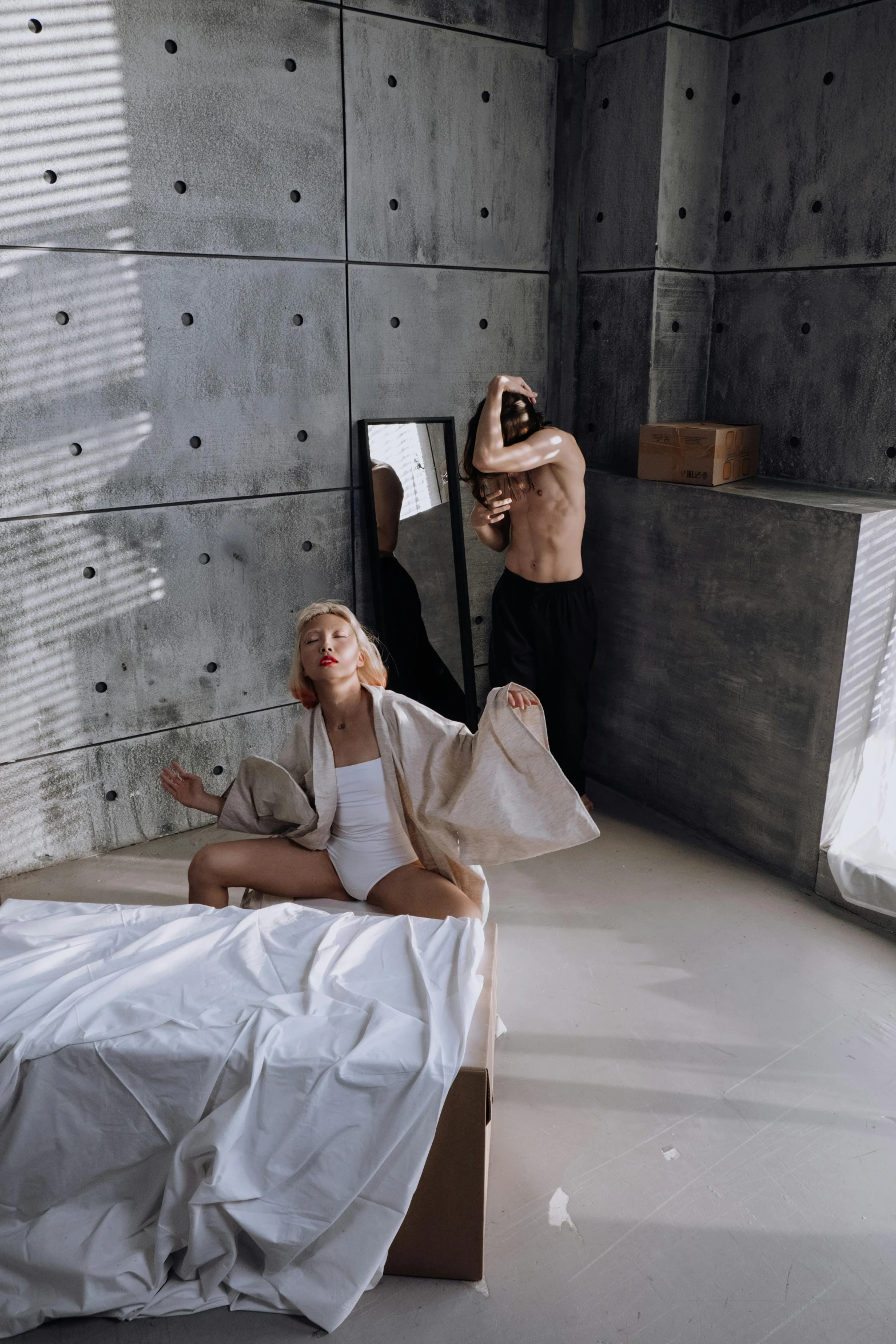 a man and woman in bedroom in cement structure with bed, mirrors, lamps