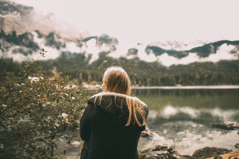 a girl sitting near a lake gazing at mountains