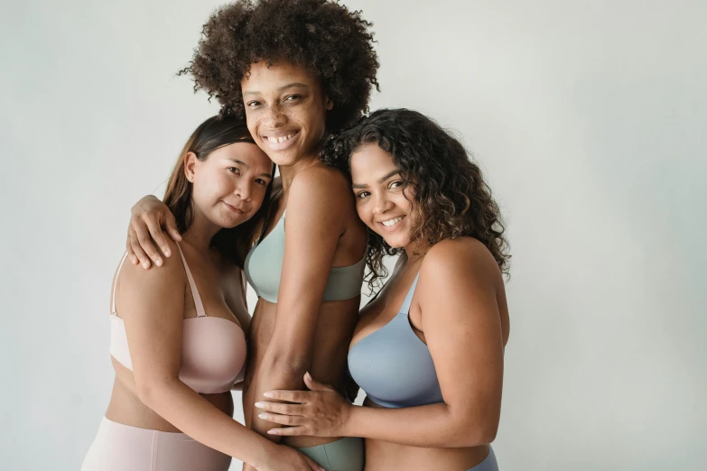 three women are hugging with each other while one woman smiles and is wearing underwear