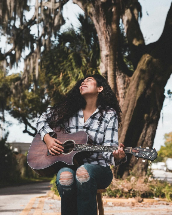 a woman with a guitar is sitting in front of a tree