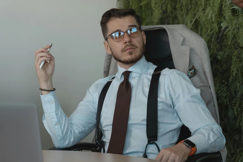 a man sitting at a desk with his legs crossed, smoking and looking up, and wearing glasses