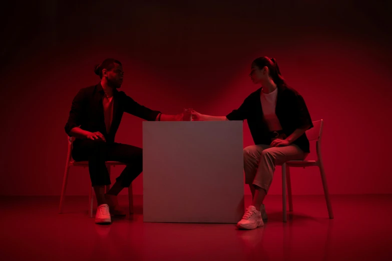 two women in black jackets sitting on white chairs and facing off in red lights