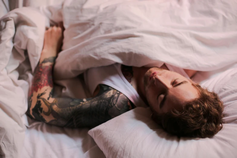 tattooed man laying on bed under blanket with white sheets