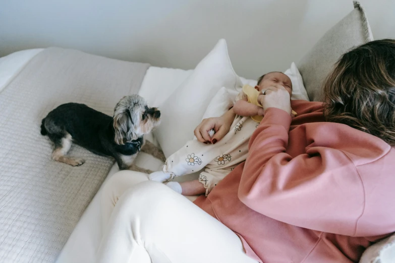 a woman in pink sits on a white bed with her baby and dog