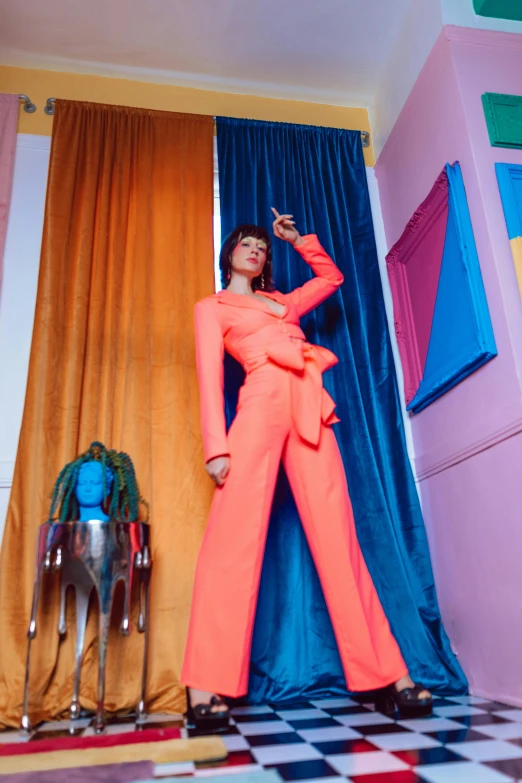 a person in an orange jumpsuit and black heels stands against a colored wall