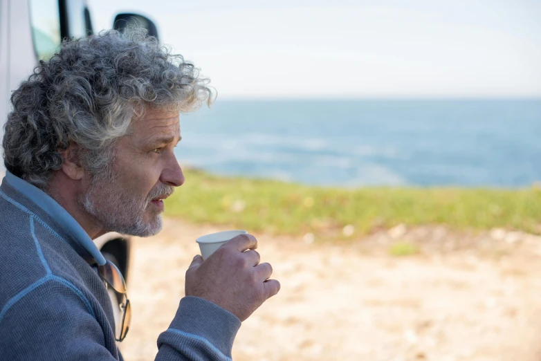 a man holding a coffee cup next to a vehicle on a dirt road