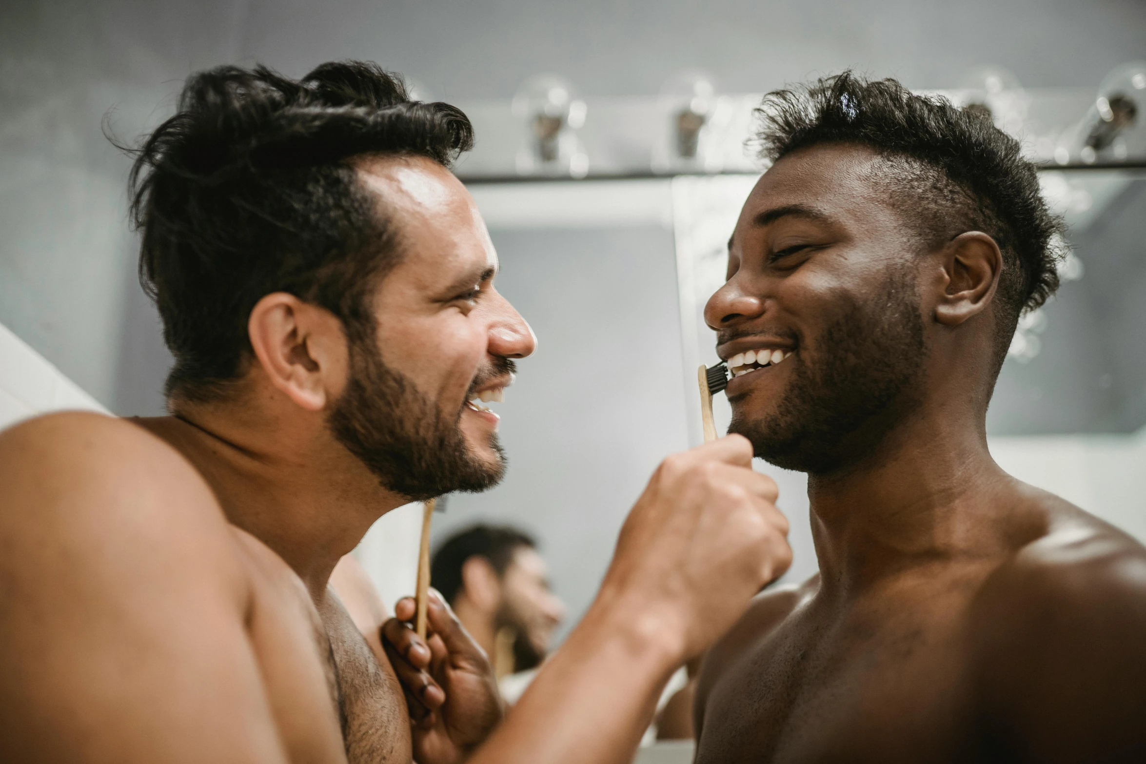 two shirtless men smile as they brush their teeth