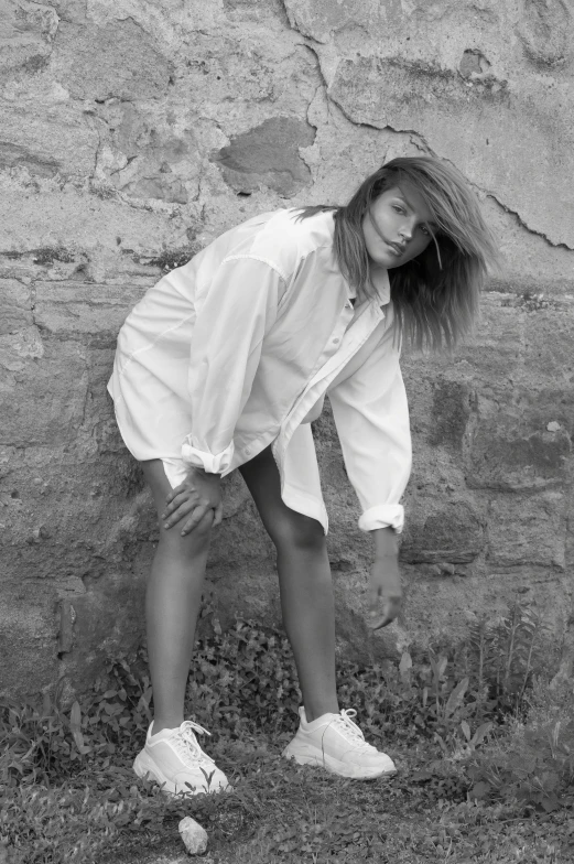 a woman in white shirt and white tennis shoes leaning against brick wall