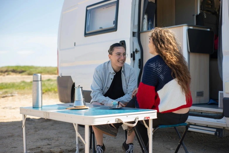 a young woman sitting at a table in front of a camper trailer with an elderly woman looking on