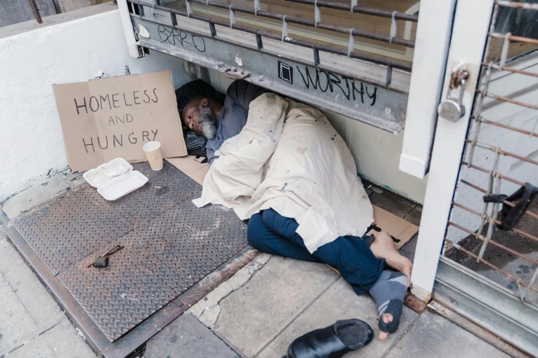homeless man lying down on the sidewalk next to a sign saying homeless you're slund on